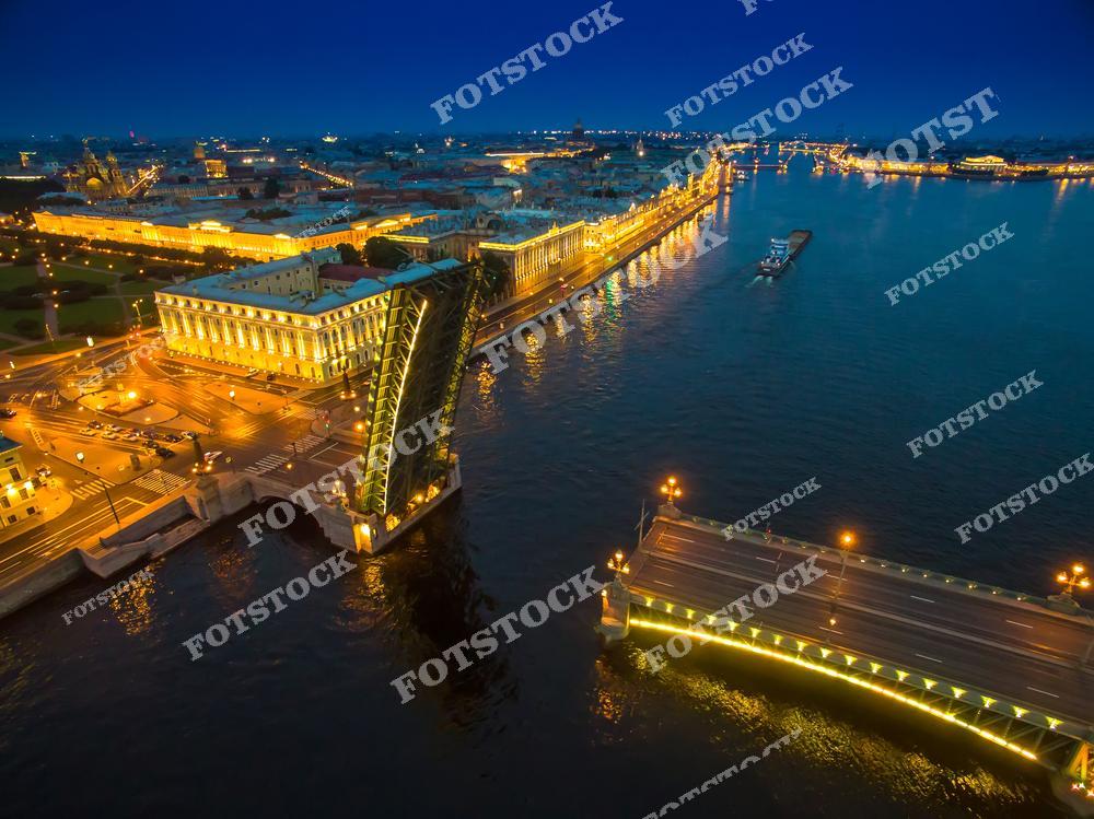 На фотографии изображен Троицкий мост в Санкт-Петербурге в момент разведения. Мост ярко освещен, а вдоль набережной Невы видны исторические здания с подсветкой, которые создают атмосферу ночного Петербурга. Троицкий мост — один из символов города, соединяющий центр Петербурга с Петроградской стороной. Разведение мостов — традиционное зрелище, привлекающее внимание туристов и жителей города.